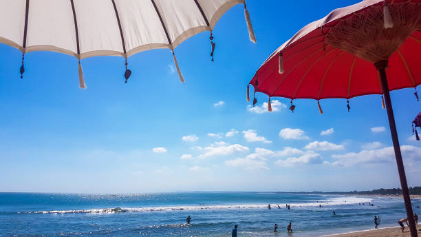 White and red linen umbrellas with decorative pendants at the ends protecting from the sunbeams on a sunny and bright day. The umbrellas are located at Seminyak Beach, Bali. Relaxation and remedy