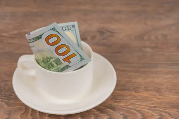 100 dollars banknote in white coffee cup on wooden table, tips or expensive service concept