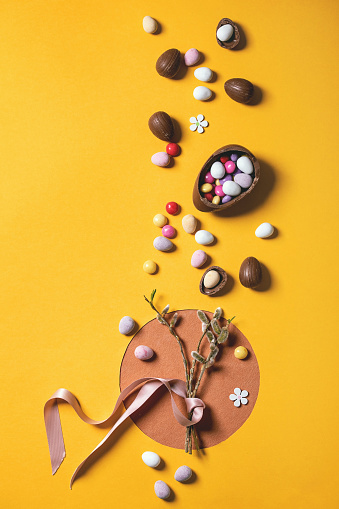 Easter greeting card with blossom willow branches, chocolate sweets and eggs on bright orange yellow background. Flat lay, copy space.