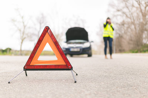 istock Young Caucasian woman, talking on her cell phone while her car is broken down on the road with reflective warning triangles. Selective focus. Automobile and Roadside Assistance Concept. 1211003724