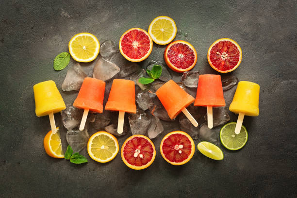 istock Assorted orange popsicles with slices of citrus, mint and ice on a dark rustic background. Frozen juice of red and yellow orange on a stick. Top view, flat lay. 1211006241