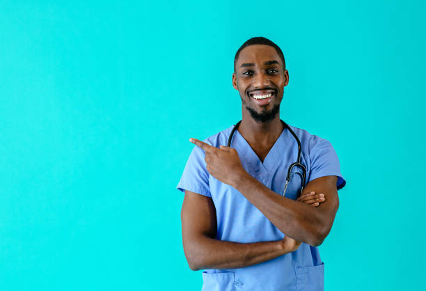 portrait of a smiling male doctor or nurse pointing with finger  to side at studio copy space and looking at camera - nurse doctor scrubs male imagens e fotografias de stock