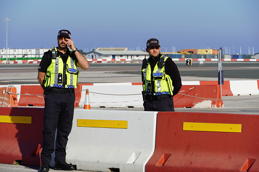 Adult hispanic male is an airport worker and is leaning on his work car. He is wearing yellow vest, blue shirt and grey work pants. He has walkie talkie pinned on pants. He is very focused. Work car has red and orange line on it and it is meant for airfield operations. Sky is blue. Focus is on airport worker.