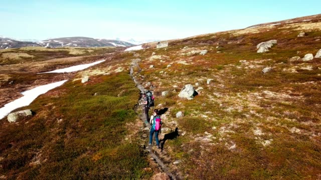 A drone flight following a couple carrying big backpack hikes through dry trail, with snow patches in few paces. High mountains in the back covered with snow. Beautiful day for a trek.