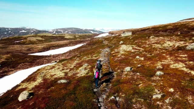 A drone flight following a couple carrying big backpack hikes through dry trail, with snow patches in few paces. High mountains in the back covered with snow. Beautiful day for a trek.