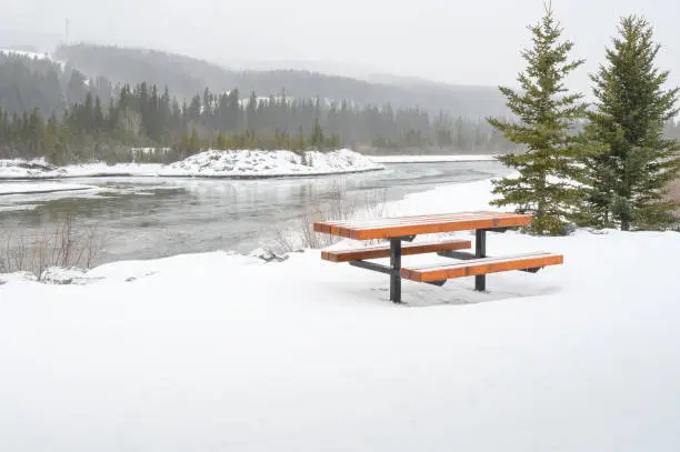 Snowy winter view of a picnic table on the bank of the Bow River in Canmore, Alberta, Canada