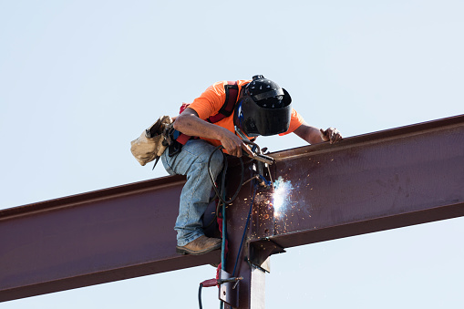 An Hispanic steel worker working high up on a girder. He is sitting on the girder, wearing a safety harness, welding to secure the girder to a column.