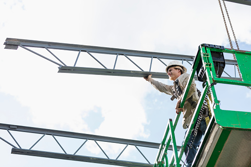 An ironworker working at a construction site, standing on a scissor lift, guiding a roof joist which is being lifted by a crane.