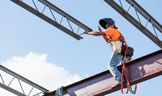An Hispanic steel worker working high up on a girder. He is standing on the girder wearing a welding helmet, reaching for a roof joist to help guide it into place.