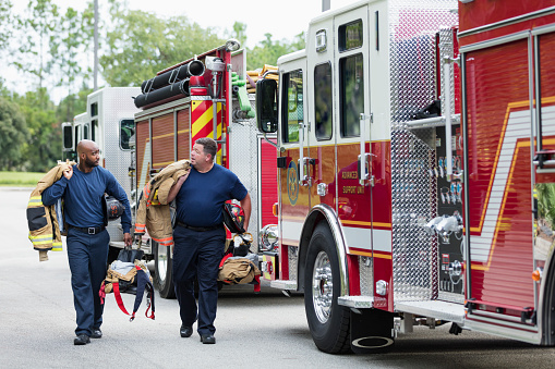 Two multi-ethnic firemen carrying equipment and fire protection suits, conversing as they walk next to two fire engines. The African-American firefighter is a young man in his 20s. His coworker is a mature heavyset man in his 40s.