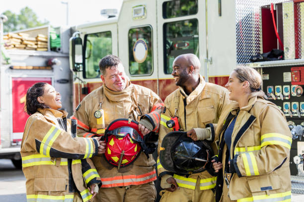 Team of firefighters standing in front of fire truck A multi-ethnic group of four firefighters standing together in front of fire engines, wearing fire protection suits and holding helmets. Two of them are women. They are smiling and conversing. firefighter stock pictures, royalty-free photos & images