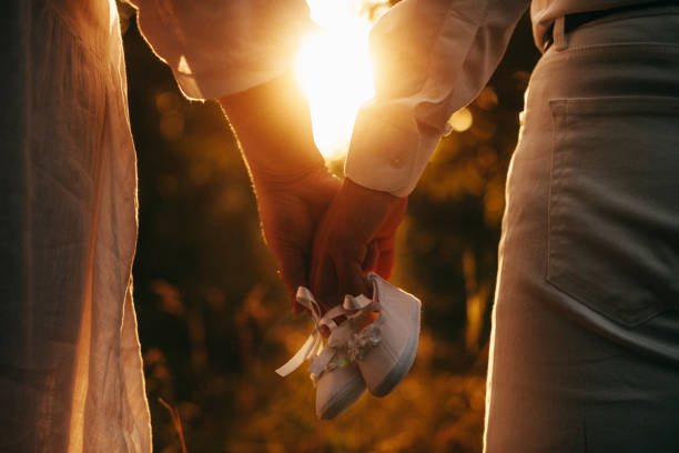 caucasian couple posing against the sunshine while walking and holding a pair of baby shoes - shoe women adult baby imagens e fotografias de stock