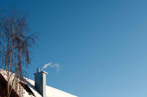 smoking chimney on a snowy roof in winter with bare birch tree beside the house