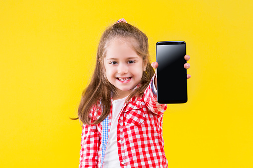 Fair haired pretty smiling girl is holding in hands smartphone with empty blank screen display. Cute child dressed in checkered pink shirt on yellow background. Modern children concept.