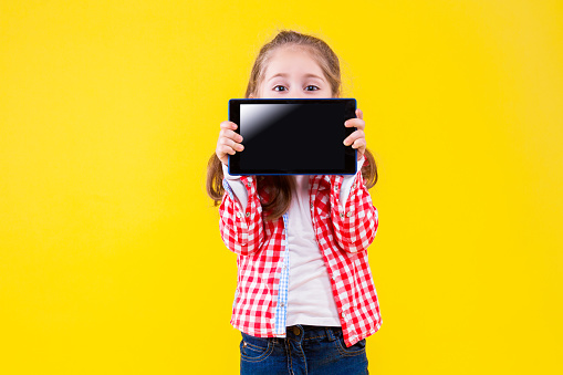 Fair haired girl is holding in hands tablet computer with empty blank screen display and hiding behind. Cute child dressed in checkered pink shirt on yellow background. Modern children concept.