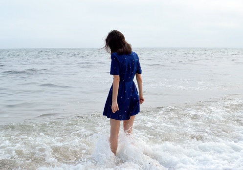 Upset thoughtful woman in blue dress going to the sea in cloudy weather before rain. She looking into the water