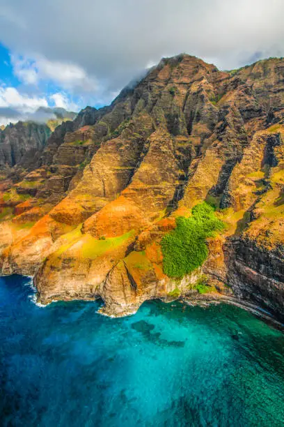 Aerial view of the famous and unique landscape of Na Pali Coast at the island of Kauai, Hawaii, on a sunny day with dramatic clouds over crystal clear water of the pacific ocean and green rocks of the island's coast