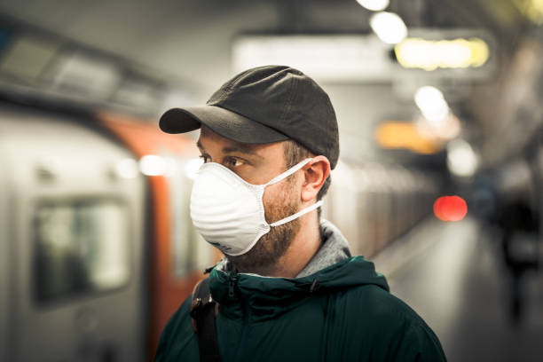 Man wearing protective face mask in city subway station Close up color image depicting a young caucasian man in his 30s wearing a white protective face mask - to protect himself from flu viruses and the coronavirus - in an underground subway station in the city of London. He is wearing casual clothing, a green rainmack and olive green baseball cap, and is looking directly at the camera. In the background, a passenger train and people are defocused. Room for copy space. london england rush hour underground train stock pictures, royalty-free photos & images