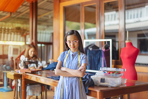 asian little girl dressmaker keeping arms crossed and looking at camera with smile while standing in workshop