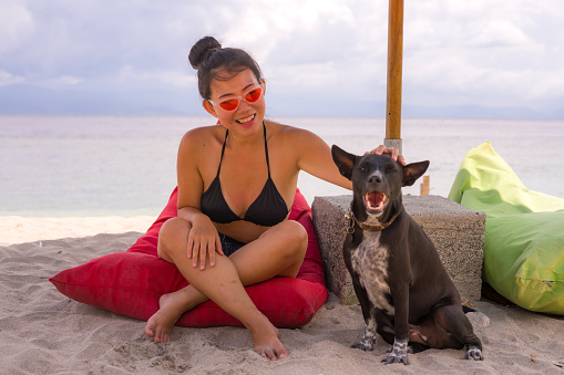 outdoors portrait of young happy and attractive Asian Chinese woman in bikini at beautiful beach relaxed and cheerful together with her dog enjoying summer holidays by the sea
