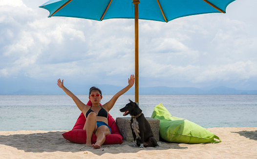 outdoors portrait of young happy and attractive Asian Korean woman in bikini at beautiful beach relaxed and cheerful together with her dog enjoying summer holidays by the sea