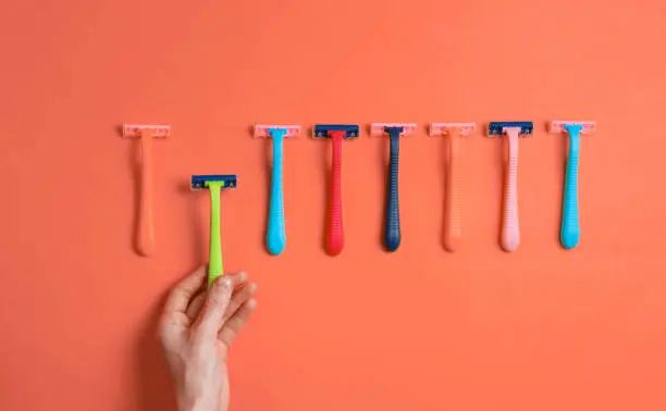 Female hand choosing green razor among a variety of colored epilator razors on orange background. Top view. Minimalism beauty concept. Top view
