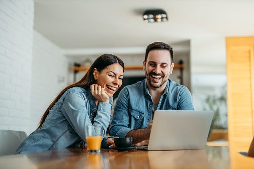 Portrait of a couple sitting at home laughing and looking at laptop screen. Dressed in casual clothes.