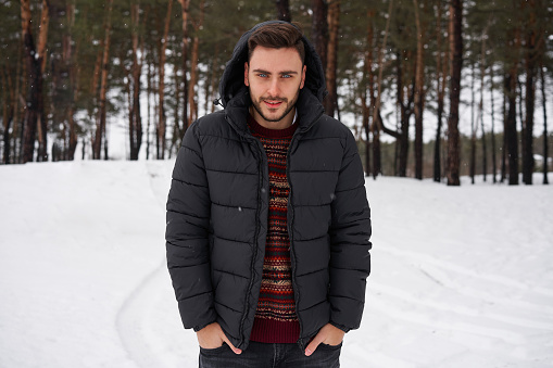 Handsome bearded young caucasian man standing outdoors in winter season forest. Attractive stylish european guy walking snowy christmas woodland. Season holiday leisure