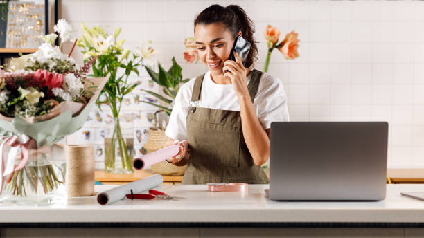 fioraio femminile in piedi nel negozio di fiori. giovane donna che parla al cellulare. - fioraio negoziante foto e immagini stock