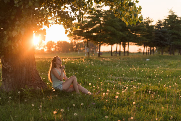 istock Woman blowing dandelion in the wind, sitting under tree in sunlight 1211212438