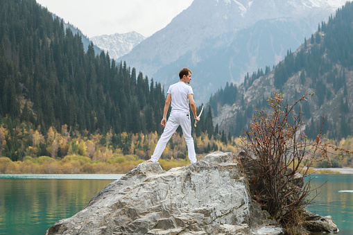 A man in white clothes with a sword stands on a large stone among the highlands.