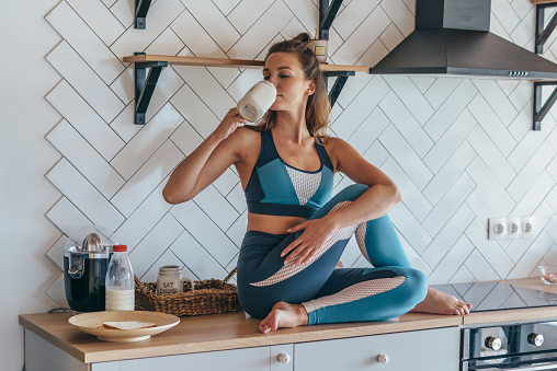 Athletic woman having breakfast in the kitchen, girl sitting on table and drinking.