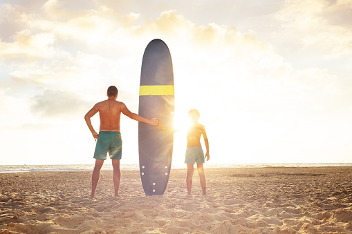 Sunset rays flare and father with son stand holding surf board on the sea beach