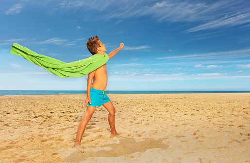 Little boy pretend to be superhero using beach towel standing on the sand and swimsuit near sea