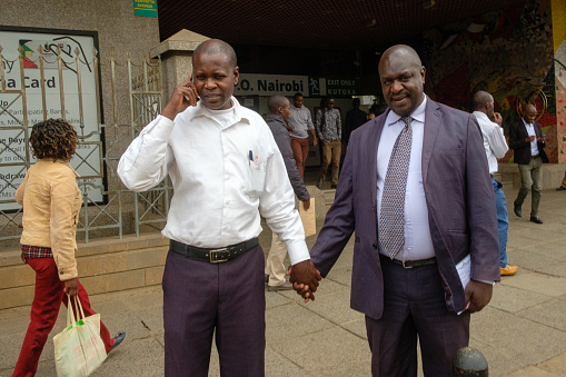 Nairobi, Kenya - August 23, 2018: Two men dressed in business clothes hold hands, as one talks on the phone, in downtown Nairobi.