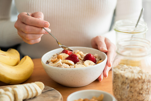 Woman eating oatmeal porridge with banana, strawberries and nuts. Healthy breakfast at the sunny morning kitchen table