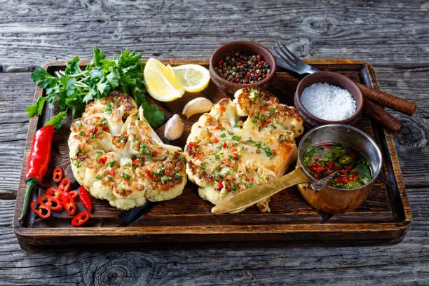 Plant-based recipe of cauliflower steaks on a wooden cutting board sprinkled with the salsa of chili, red vinegar, garlic, and cilantro, served with lemon and cutlery on a wooden table, close-up