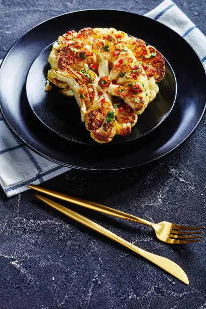 Delicious oven baked cauliflower steaks on a black plate sprinkled with the salsa of red chili pepper, vinegar, garlic, and cilantro, served with golden cutlery, vertical orientation, close-up