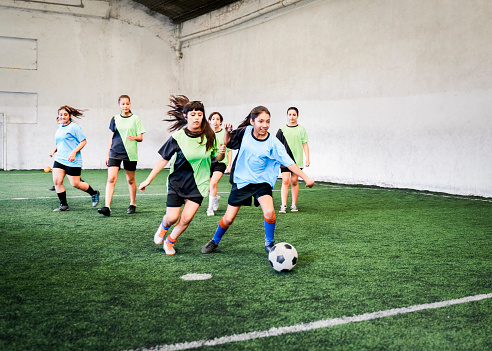 Female players in blue and green jerseys playing soccer. Girls are running towards ball during match. They are at sports court.