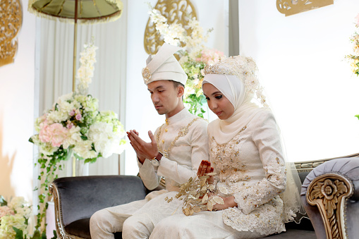 A pair of newlywed Muslim bride and groom seated for prayers at 'pelamin' (a decorated newlywed seating at the main stage) in wedding reception, Kuala Lumpur Malaysia.