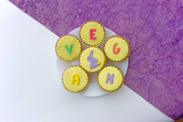 istock Pretty yellow vegan Easter cupcakes with icing shapes on plate, on two tone white and purple textured background, top view 1211287625