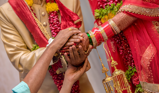 Traditional Indian Wedding Photo.\nConcept of Kanyadaan, Ritual, Customs, Hindu Marriage, Vedic Marriage, Relationships, Husband Wife, Love Marriage, Arrange Marriage, Matrimony, Match making, Dowry marriage etc. depending on the vision of user.\nAmazing photo of a traditional Indian wedding taking place wherein the priest/pandit is holding the hands of bride and the groom together to tie them in the relationship of husband and wife.\n The bride and the groom are wearing ethnic Indian attire and are offering prasadam to fire god to receive blessings for their happy married life. Bride alongwith the dress is also wearing colorful bridal bangles and floral garland is worn by both bride and groom.