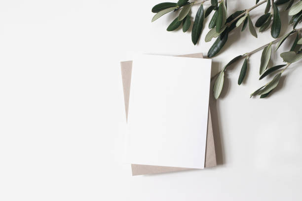 istock Summer wedding stationery mock-up scene. Blank vertical greeting card, craft paper envelope and olive branches isolated on white table background. Feminine Mediterranean flat lay, top view. 1211298144