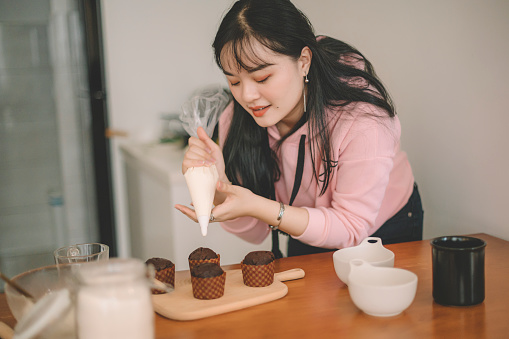 an asian chinese young female putting icing on cupcake in the kitchen