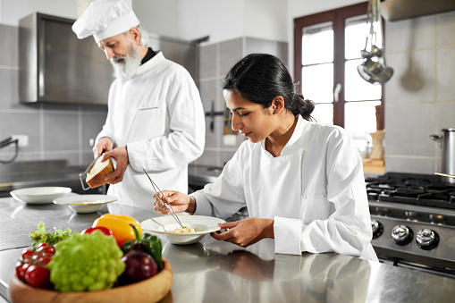 Female indian sous chef is plating her dish in the kitchen with tweezers. She is focused and confident. Behind her is the head male chef preparing their next step for their guests in the restaurant. They are wearing chefs uniforms in white color.