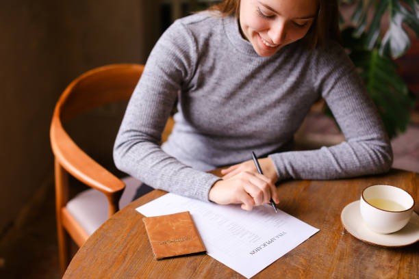 istock Young woman filling visa application at cafe, passport on table 1211307310