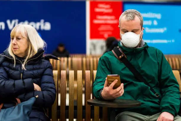 Close up color image depicting a young caucasian man in his 30s wearing a white protective face mask - to protect himself from flu viruses and the coronavirus - sitting and waiting on a bench at a railroad station in the city of London. The man is sitting next to a senior woman in her 60s or 70s, and she is not wearing a face mask. The man is wearing casual clothing, a green rainmack. He is looking at his smart phone. In the background other people are defocused.