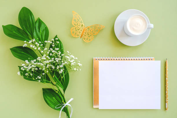 Workspace with blank sheet of notepad, pencil, green leaf, decorative golden butterfly and coffee cup on green background. Mock up Top view Flat lay Template for card, design
