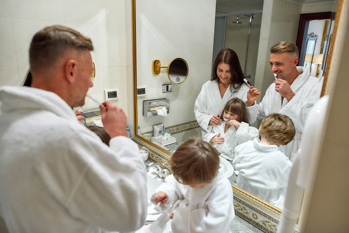 Caucasian parents and two kids in white bathrobes having morning routine in hotel bathroom, standing near the mirror and brushing teeth. Family vacation concept. Horizontal shot