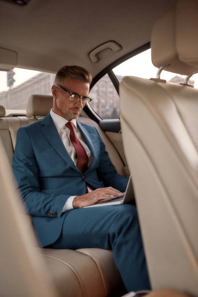 istock Never stop working. Serious mature businessman in full suit working on his laptop while sitting in the car 1211311817